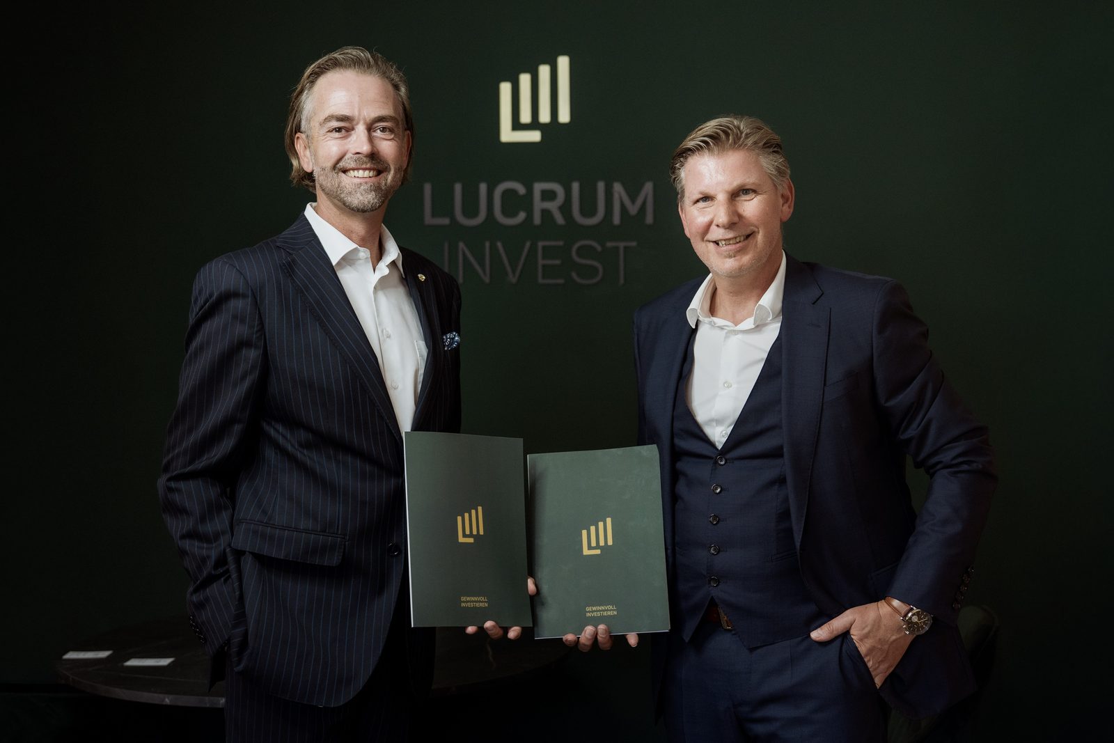Roman Zlabinger und Helmut Hemmelmeir mit Beratungsmappen vor der Logowand von Lucrum Invest