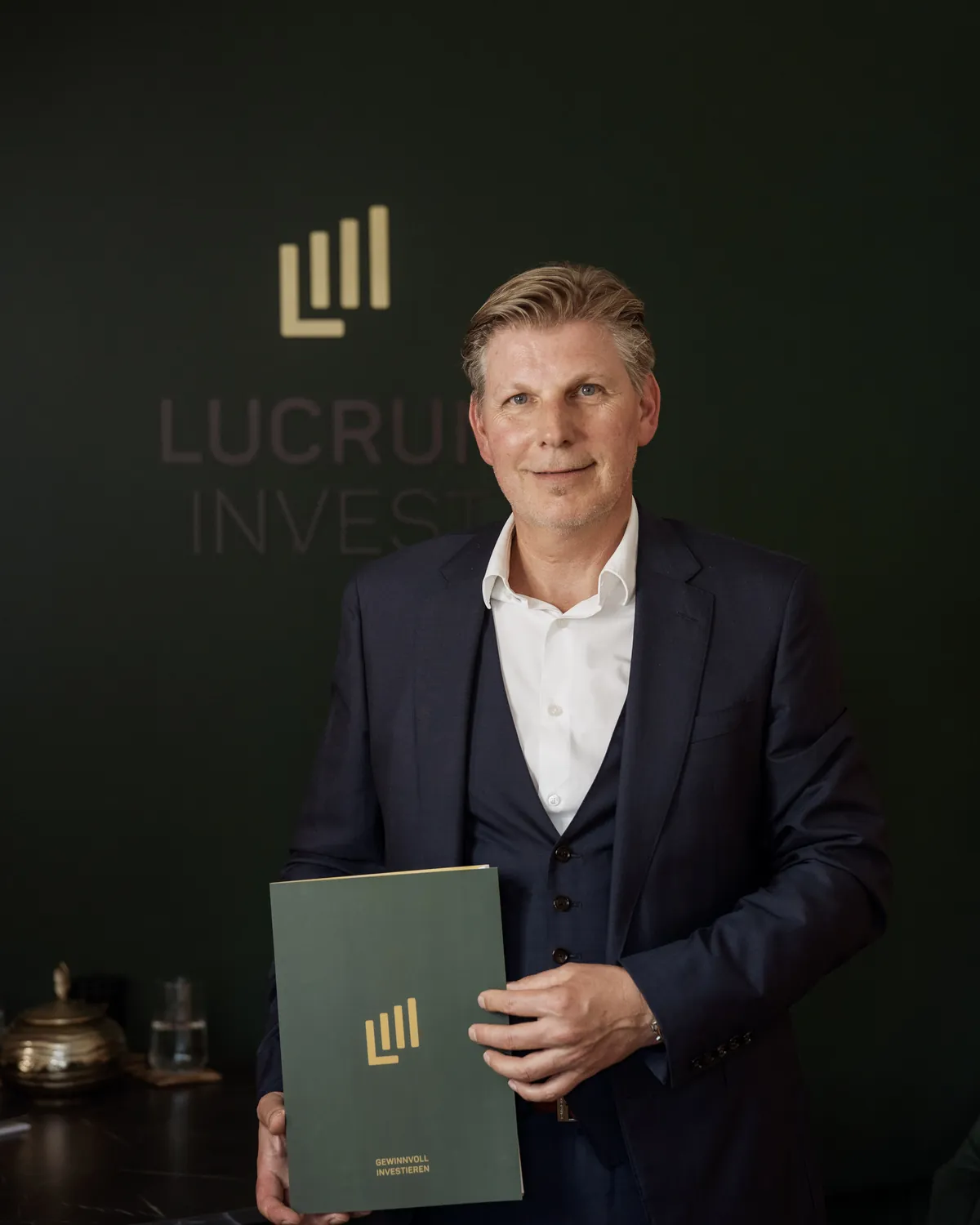Helmut Hemmelmeir mit einer Beratungsmappe vor der Logowand von Lucrum Invest