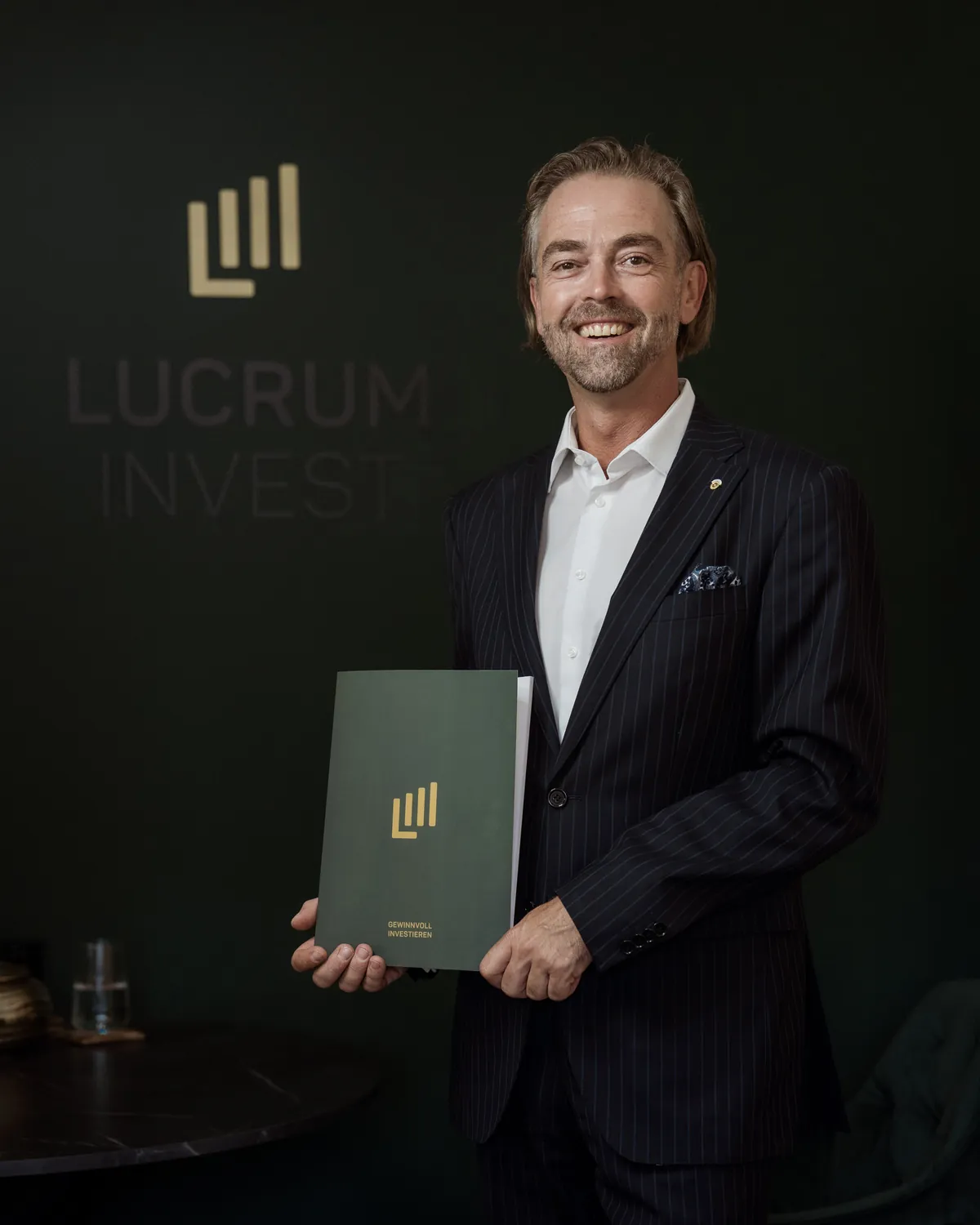 Roman Zlabinger mit einer Beratungsmappe vor der Logowand von Lucrum Invest