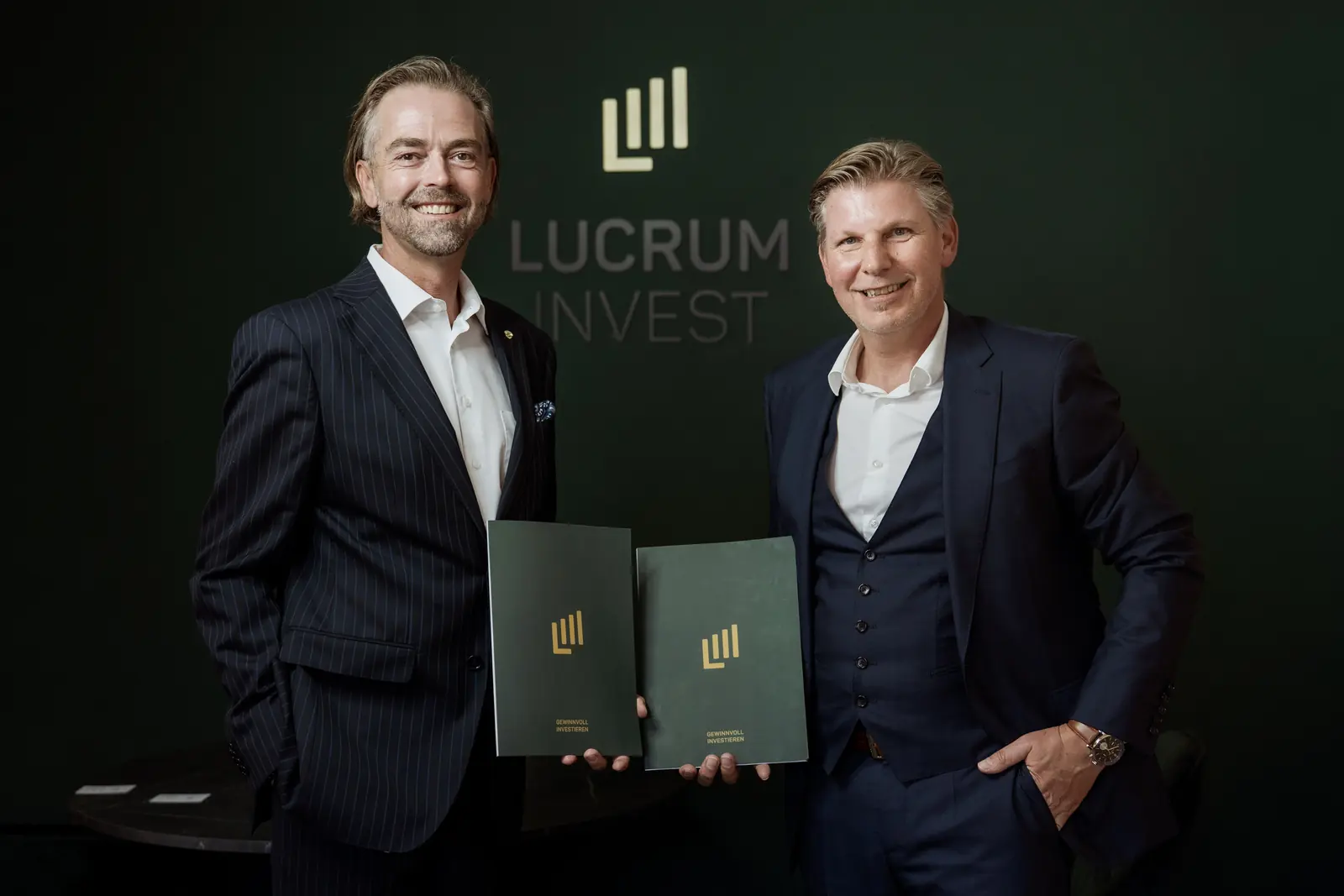 Roman Zlabinger und Helmut Hemmelmeir mit Beratungsmappen vor der Logowand von Lucrum Invest
